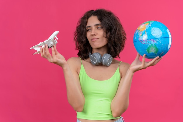 A young attractive woman with short hair in green crop top in headphones holding globe and looking at toy plane