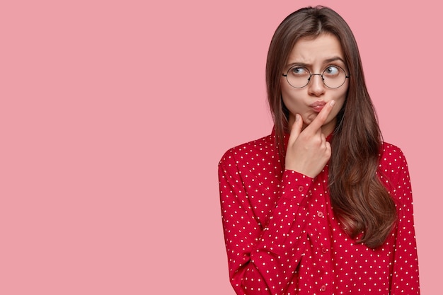 Free photo young attractive woman with thoughtful expression, thinks about question, feels doubt, raises eyebrows, holds chin