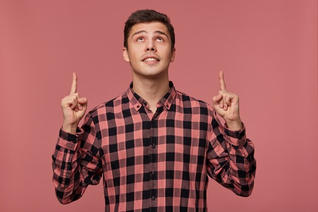 Young attractive wondered man wears in checkered shirt, looks up and wants to turn your attention to copy space over his head , isolated over pink background