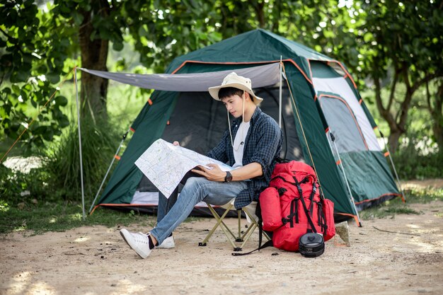 모자를 쓴 젊은 배낭 여행자는 자연 숲의 텐트 앞에 앉아 숲길의 종이 지도를 보고 여름 방학 동안 캠핑 여행을 계획하고 공간을 복사합니다.