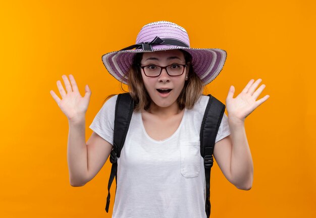 Молодая женщина-туристка в белой футболке в летней шляпе с рюкзаком весело улыбается, глядя удивленно, стоя над оранжевой стеной