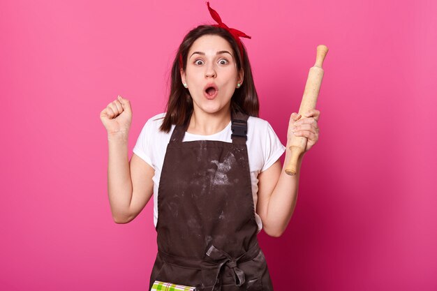 Young baker on pink wall looks at camera, bends elbows, holds rolling pin. Charming lady with happy facial expression has new idea while baking chocolate cookies. Food and culinary concept.