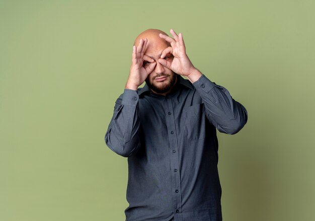 Young bald call center man doing look gesture at camera using hands as binoculars isolated on olive green background with copy space