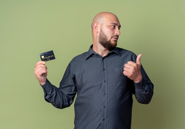 Young bald call center man holding credit card showing thumb up and looking at side isolated on olive green background