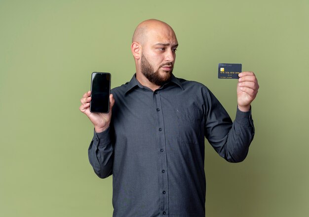 Young bald call center man holding mobile phone and credit card and looking at card isolated on olive green background with copy space
