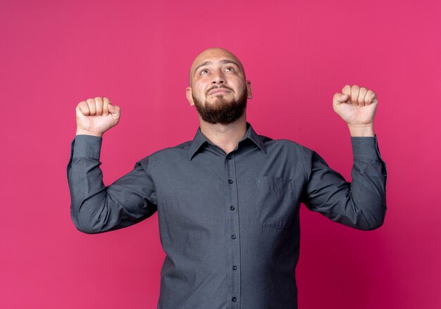 Young bald call center man raising fists and looking up isolated on crimson background