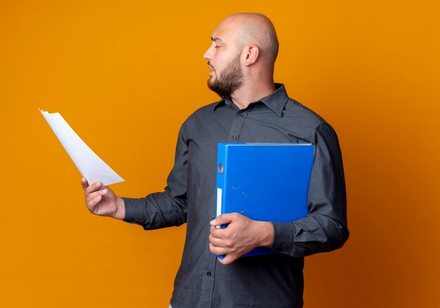 Young bald call center man turning head to side with closed eyes holding folder and document isolated on orange background