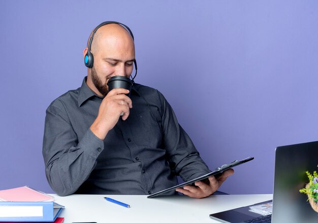 Young bald call center man wearing headset sitting at desk with work tools holding and looking at clipboard and drinking coffee isolated on purple background