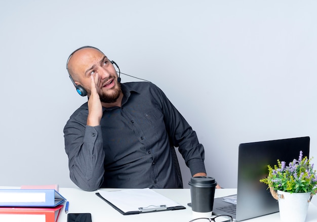 Young bald call center man wearing headset sitting at desk with work tools looking up calling out to someone isolated on white background