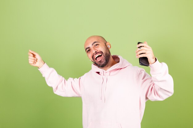 Young bald caucasian man in pink hoodie isolated, hold phone happy dancing moving in headphones enjoying with closed eyes