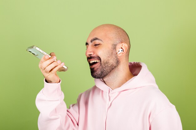 Young bald caucasian man in pink hoodie isolated, hold phone happy positive recording audio message