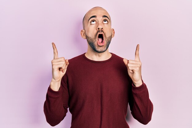 Young bald man wearing casual clothes amazed and surprised looking up and pointing with fingers and raised arms.