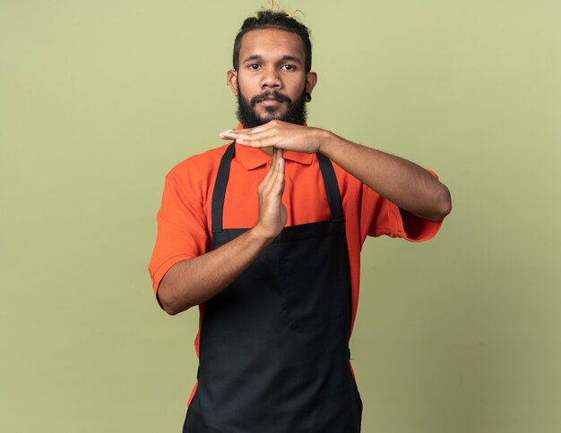 Young barber wearing uniform looking at front doing timeout gesture isolated on olive green wall with copy space