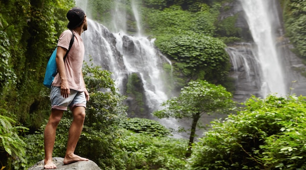 Молодой босоногий турист в бейсболке стоит на большом камне и смотрит на водопад позади него в красивой экзотической природе. Бородатый путешественник наслаждается дикой природой во время похода в тропический лес