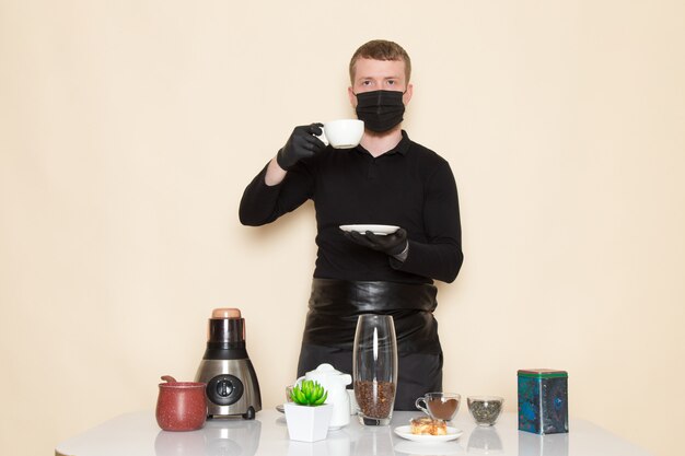 young barista in black working suit with ingredients and coffee equipment brown coffee seeds wearing black sterile mask on white