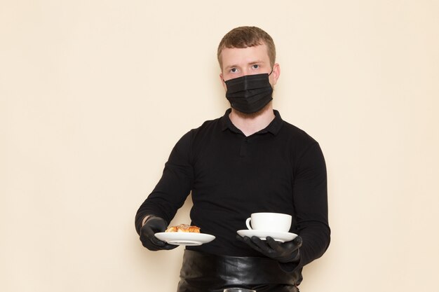 молодой бариста в черном рабочем костюме с ингредиентами и кофейным оборудованием коричневые семена кофе в черной стерильной маске на белом