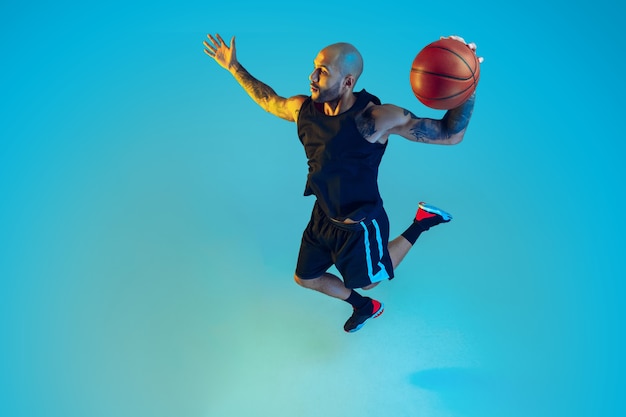 Young basketball player of team wearing sportwear training, practicing in action, motion on blue wall in neon light