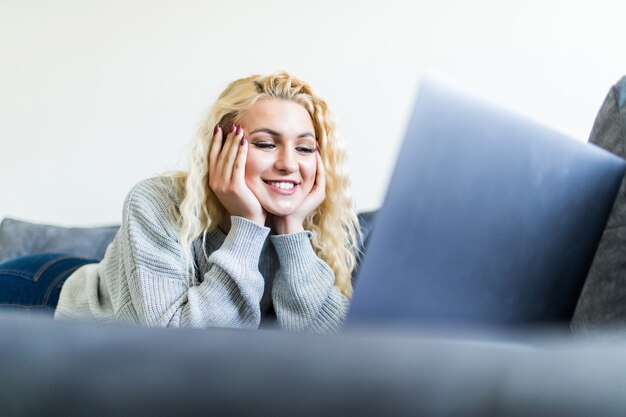 Young beaauty woman lying on couch working on laptop in apartment