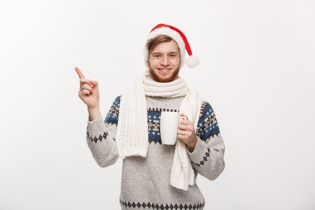 흰색 측면에 손을 가리키는 뜨거운 커피 컵을 들고 스웨터와 산타 모자에 젊은 수염 남자