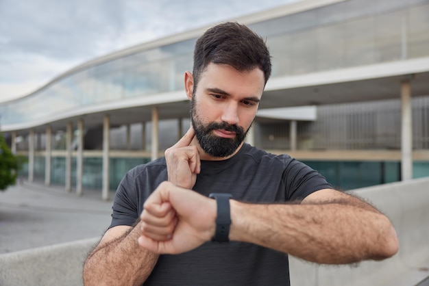 Young bearded adult man checks pulse using smartwatch dressed in casual black t shirt poses against urban background measures heart rate during workout monitors performance after exercising.