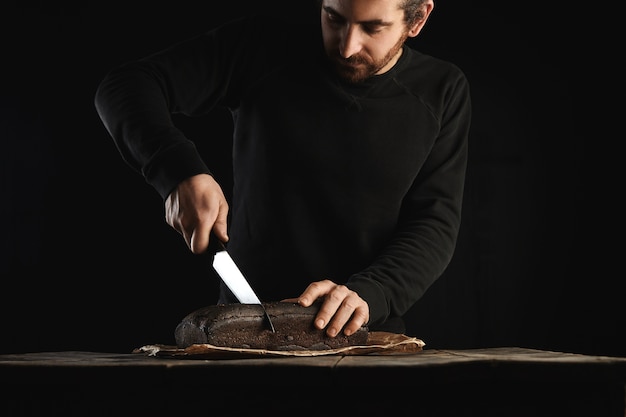 Young bearded baker man in black sweatshot uses big chief knife to slice homemade luxury bread from figs and rye in craft paper on rustic wooden table isolated on black