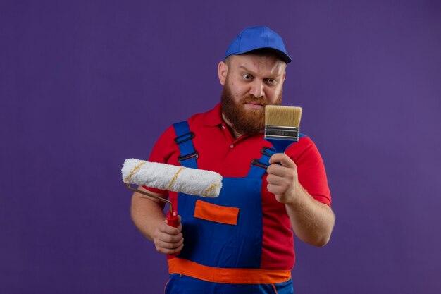 Young bearded builder man in construction uniform and cap holding paint roller and brush looking at brush with skeptic expression on face over purple background