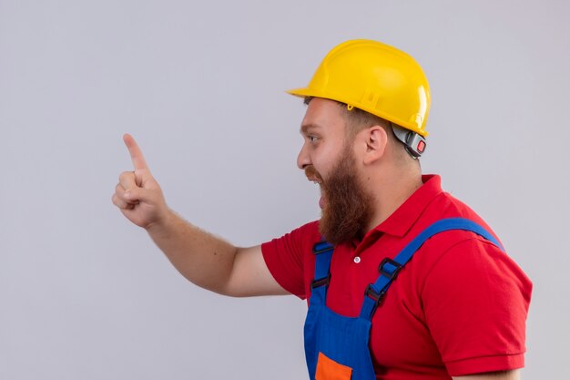 Молодой бородатый строитель в строительной форме и защитном шлеме эмоционально и взволнованно кричит, показывая указательный палец