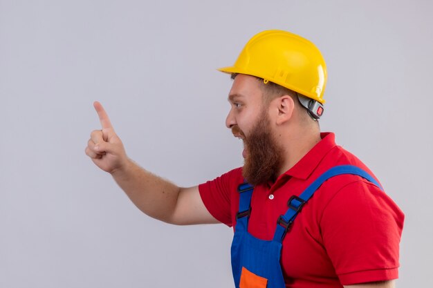 Young bearded builder man in construction uniform and safety helmet emotional and worried shouting showing index finger