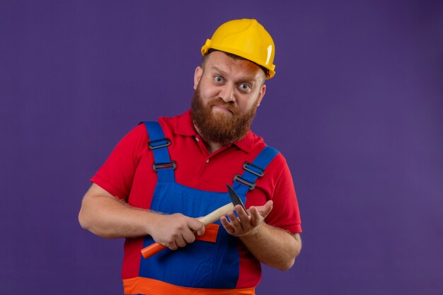 Young bearded builder man in construction uniform and safety helmet holding hammer with frowning face over purple background