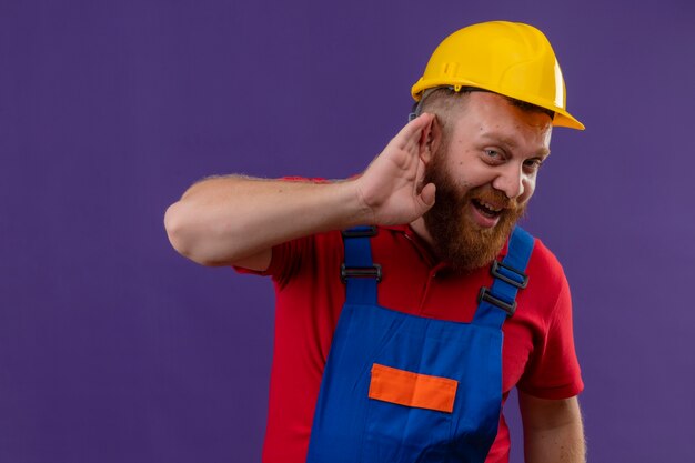 Young bearded builder man in construction uniform and safety helmet holding hand near his ear trying to listen someone conversation over purple background