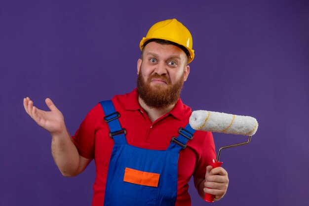 Молодой бородатый строитель в строительной форме и защитном шлеме, держащий валик с краской, выглядит смущенным, пожимает плечами и сомневается на фиолетовом фоне