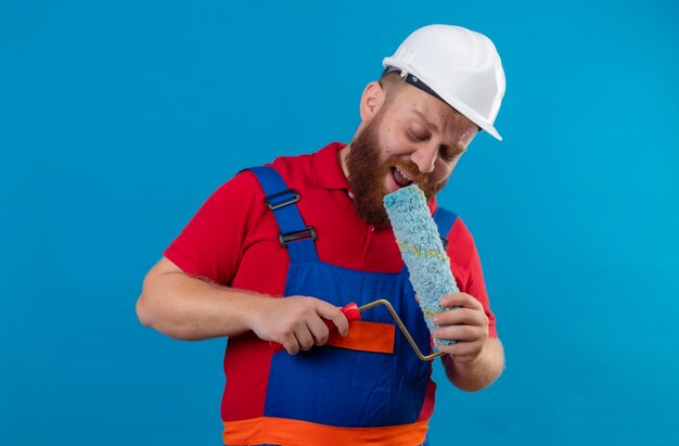 Young bearded builder man in construction uniform and safety helmet holding paint roller using as a microphone singing, having fun at work 
