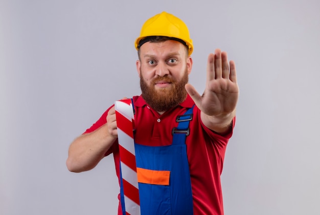 Young bearded builder man in construction uniform and safety helmet holding scotch tape standing with open hand making stop sign over white background