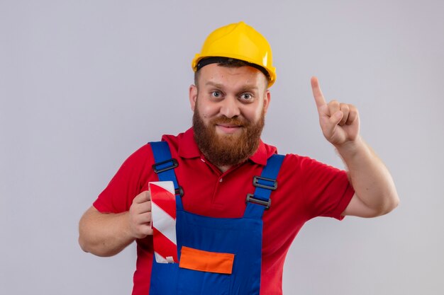 젊은 수염 작성기 남자 건설 유니폼 및 안전 헬멧 들고 테이프 스카치 카메라를보고 웃 고 가리키는 손가락