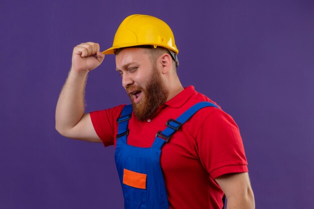 Молодой бородатый строитель в строительной форме и защитном шлеме раздраженно сжимает кулак на фиолетовом фоне