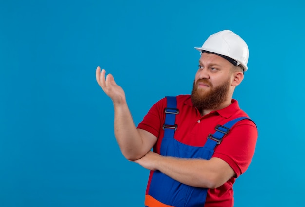 Молодой бородатый строитель в строительной форме и защитном шлеме недовольно смотрит в сторону с поднятой рукой