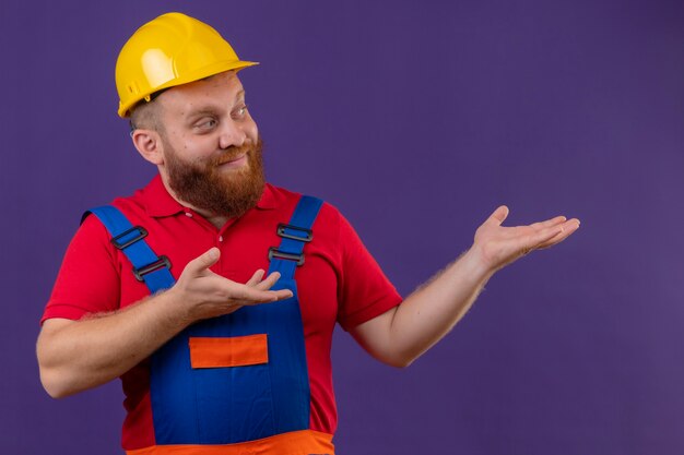 Молодой бородатый строитель в строительной форме и защитном шлеме смотрит в сторону, демонстрируя руки на фиолетовом фоне