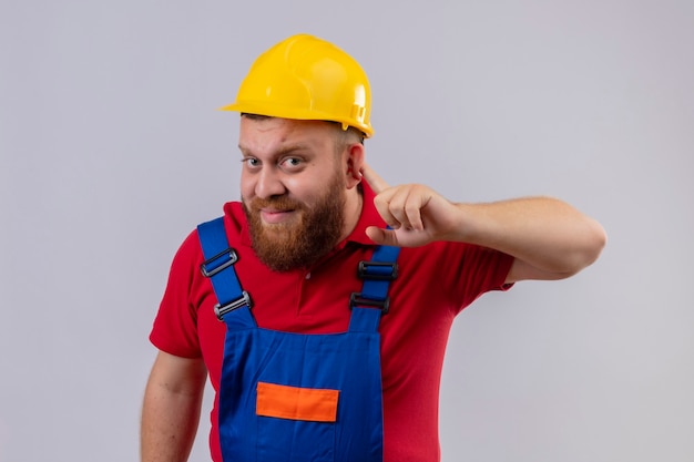 耳の近くで手をつないでカメラを見て聞いている建設制服と安全ヘルメットの若いひげを生やしたビルダーの男