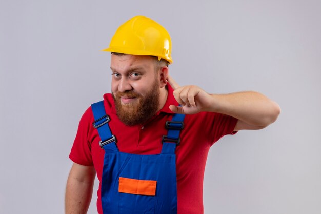 Young bearded builder man in construction uniform and safety helmet looking at camera holding hand near ear trying to listen