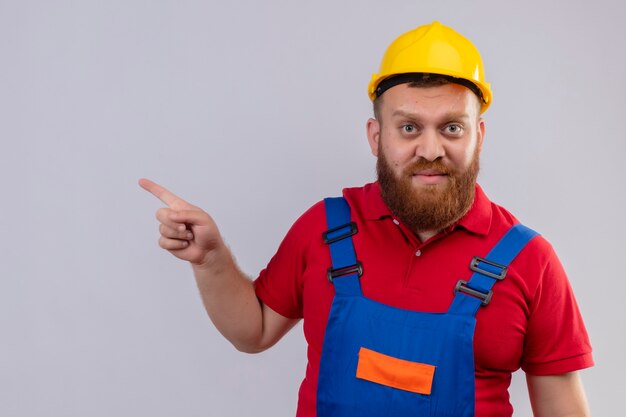 Young bearded builder man in construction uniform and safety helmet looking at camera pointing with finger to the side with serious face