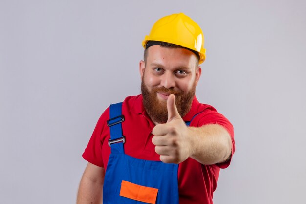 Young bearded builder man in construction uniform and safety helmet looking at camera smiling showing thumbs up