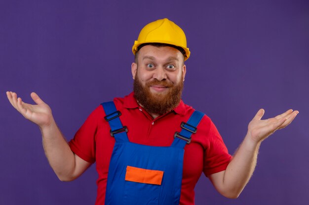 Young bearded builder man in construction uniform and safety helmet looking surprised and confused smiling spreading palms over purple background