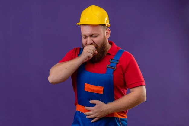 Молодой бородатый строитель в строительной форме и защитном шлеме устал, зевая на фиолетовом фоне