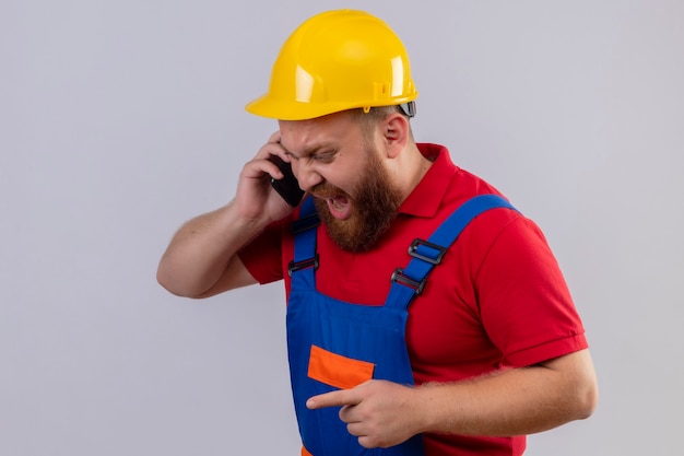 Young bearded builder man in construction uniform and safety helmet shouting while talking on mobile phone with aggressive expression