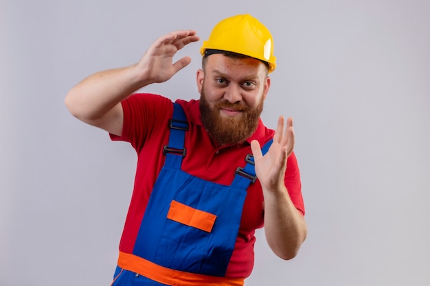 Young bearded builder man in construction uniform and safety helmet showing size gesture with hands with confident smile on face, measure symbol