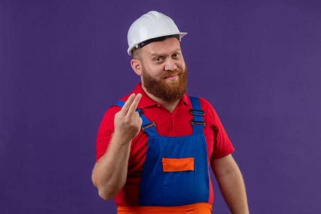 Young bearded builder man in construction uniform and safety helmet showing two fingers looking displeased over purple background