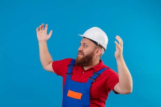 Young bearded builder man in construction uniform and safety helmet shrugging shoulders, looking confused and uncertain , having no answer, spreading palms2