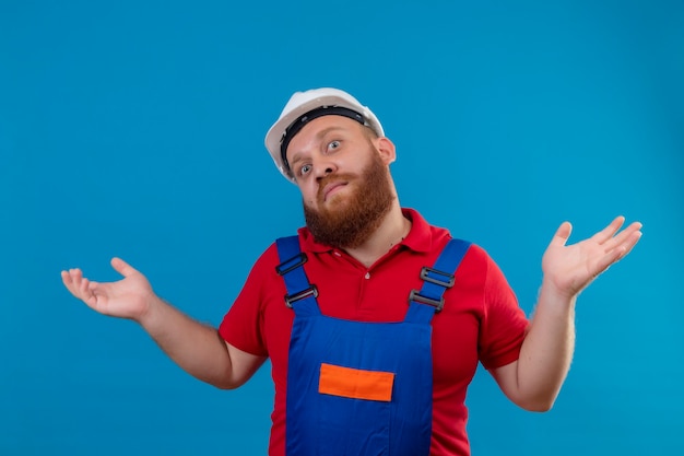 Free photo young bearded builder man in construction uniform and safety helmet shrugging shoulders, looking confused and uncertain , having no answer, spreading palms