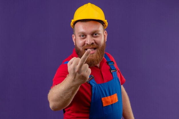 건설 유니폼 및 안전 헬멧에 젊은 수염 작성기 남자 보라색 배경 위에 가운데 손가락을 유쾌하게 보여주는 미소