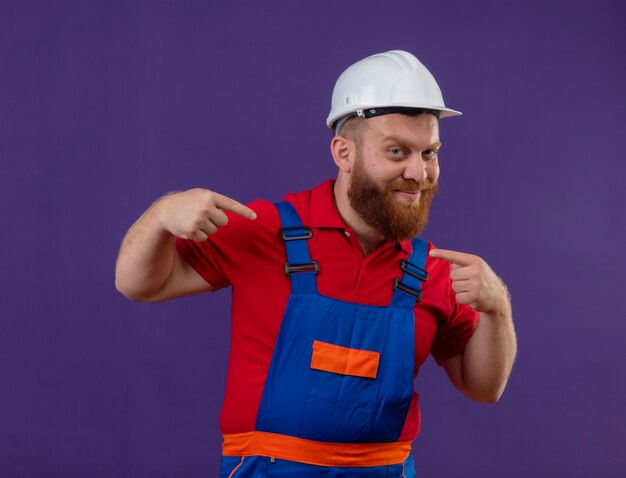 건설 유니폼 및 안전 헬멧에 젊은 수염 작성기 남자 보라색 배경 위에 검지 손가락으로 자신을 가리키는 자신감 미소